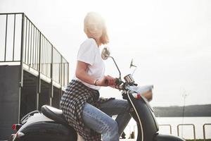 Retrato de una hermosa chica hipster sentada en un scooter retro negro, sonriendo posando y disfrutando del cálido sol primaveral. foto