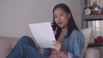 asiatisk kvinna som pratar i telefon och läser dokument medan hon sitter i soffan i vardagsrummet hemma. video