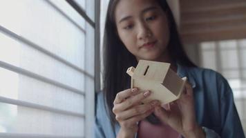 Young Asian woman freelance architect holding model of home standing beside window at home office. video