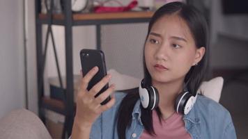 Asian woman having video chat using smartphone while sitting sofa in living room at home.