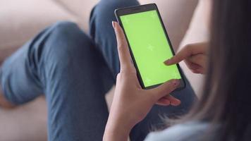 aziatische vrouw die groen scherm bekijkt op mobiele telefoon die op sociale media surft terwijl ze thuis op de bank in de woonkamer zit. video