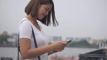 aziatische vrouw die smartphone gebruikt en geniet van het lezen van sociale media naast het hek aan de rivier. video