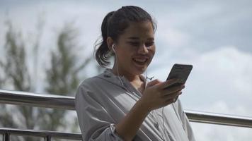 Corredor de fitness mujer asiática de pie con teléfono móvil escuchando música en un parque público. video