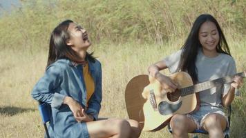 Mujer asiática feliz con amigos acampando en la naturaleza divirtiéndose juntos tocando la guitarra y bebiendo cerveza y tintineando vasos. video