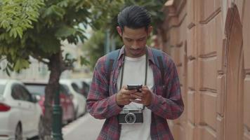 Hombre asiático con un teléfono inteligente comprobando el mapa mientras camina al lado de la calle en Tailandia. video