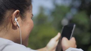 corredor de fitness mulher asiática em pé usando telefone celular ouvindo música em um parque público. video