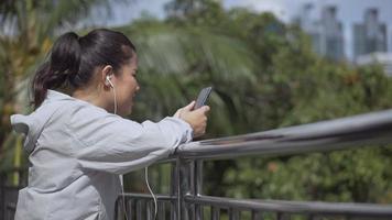 Aziatische vrouw fitness runner staande met behulp van mobiele telefoon luisteren muziek in een openbaar park. video