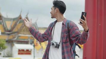 turistas solo de homem asiático em pé usando o bate-papo por vídeo de smartphones com amigos na rua, tendo o fundo do templo da Tailândia.