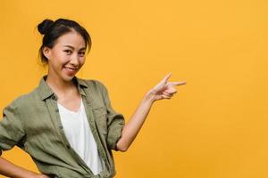 Retrato de joven asiática sonriendo con expresión alegre, muestra algo sorprendente en el espacio en blanco en un paño casual y mirando a cámara aislada sobre fondo amarillo. concepto de expresión facial.
