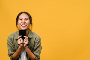 Surprised young Asia lady using mobile phone with positive expression, smile broadly, dressed in casual clothing and looking at camera on yellow background. Happy adorable glad woman rejoices success. photo