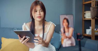 Happy young Asian girl blogger in front of phone camera use tablet enjoy question answer with follower in living room at home. Blogger activity lifestyle, Social distance coronavirus pandemic concept.