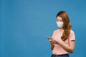 Young Asia girl wearing medical face mask using mobile phone with dressed in casual clothing isolated on blue background. Self-isolation, social distancing, quarantine for corona virus prevention. photo