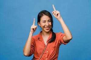 Portrait of young Asian lady smiling with cheerful expression, shows something amazing at blank space in casual cloth and looking at camera isolated over blue background. Facial expression concept. photo