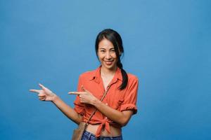 Portrait of young Asian lady smiling with cheerful expression, shows something amazing at blank space in casual cloth and looking at camera isolated over blue background. Facial expression concept. photo