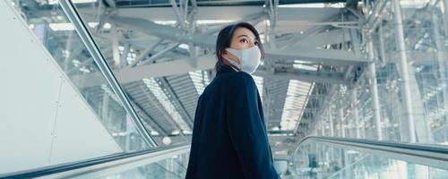 chica de negocios asiática usa mascarilla arrastre soporte de equipaje en la escalera mecánica mire alrededor camine hasta la terminal en el aeropuerto internacional. concepto de distanciamiento social de viajes de negocios. Fondo de banner panorámico. foto