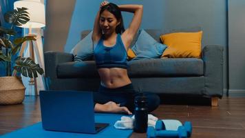 Young Asia lady in sportswear exercises doing work out and using laptop to watch yoga video tutorial at home night. Distant training with personal trainer, social distance, online education concept. photo