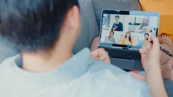 Young Asia businessman using laptop talk to colleagues about plan in video call meeting while work from home at living room. Self-isolation, social distancing, quarantine for corona virus prevention. photo