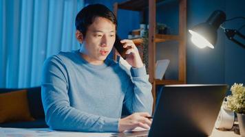 Young Asia businessman use smartphone call meeting agenda assignment paperwork with colleague look at laptop computer in living room at home overtime at night, Work from home corona pandemic concept. photo