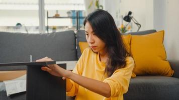 Happy asian young woman unpacking box and reading the instructions to assemble new furniture decorate house build table with carton box in living room at home. photo