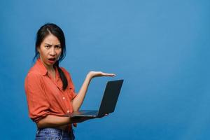 Young Asia lady using laptop with negative expression, excited screaming, cry emotional angry in casual cloth and stand isolated on blue background with blank copy space. Facial expression concept. photo