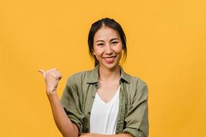 Retrato de joven asiática sonriendo con expresión alegre, muestra algo sorprendente en el espacio en blanco en un paño casual y mirando a cámara aislada sobre fondo amarillo. concepto de expresión facial.