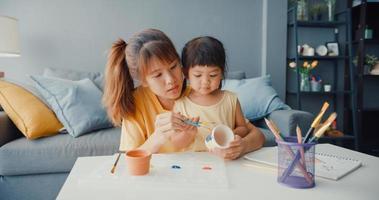 Happy cheerful Asia family mom teach toddler girl paint ceramic pot having fun relax on table in living room at house. Spending time together, Social distance, Quarantine for coronavirus prevention. photo
