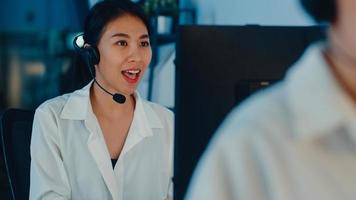 Millennial Asia young call center team or customer support service executive using computer and microphone headset working technical support in late night office. Telemarketing or sales job concept. photo