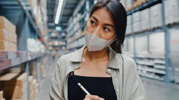 Young Asia businesswoman manager wear face mask looking for goods using digital tablet checking inventory levels stand in retail shopping center. Distribution, Logistics, Packages ready for shipment. photo