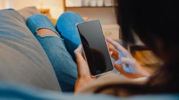 Joven mujer asiática usa un teléfono inteligente con pantalla negra en blanco simulada para mostrar texto publicitario mientras descansa en el sofá de la sala de estar en la moderna noche de casa. tecnología chroma key, concepto de diseño de marketing. foto