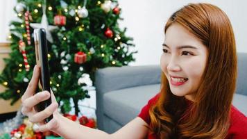 Joven mujer asiática con videollamada de teléfono inteligente hablando con pareja con caja de regalo de Navidad, árbol de Navidad decorado con adornos en la sala de estar en casa. festival de navidad y año nuevo. foto