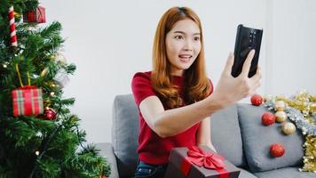 Young Asian female using smart phone video call talking with couple with X'Mas present box, Christmas tree decorated with ornament in living room at home. Christmas and New Year holiday festival. photo