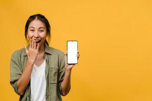Joven asiática muestra la pantalla vacía del teléfono inteligente con expresión positiva, sonríe ampliamente, vestida con ropa casual sintiendo felicidad sobre fondo amarillo. teléfono móvil con pantalla en blanco en mano femenina.