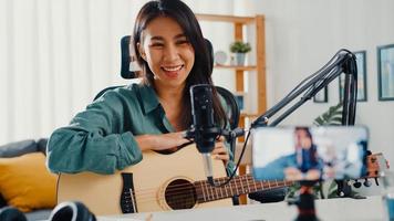 Teenage Asia girl influencer play guitar music use microphone record with smartphone for online audience listen at home. Female podcaster make audio podcast from her home studio, Stay at home concept. photo