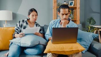 Asia young couple man and woman sit on couch serious focus on laptop computer check document paper pay bills online plan budget expense in living room. Young married asian debt loan problem concept. photo