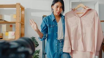 Young Asia lady fashion designer using mobile phone receiving purchase order and show clothes recording video live streaming online with camera. Small business owner, online market delivery concept. photo