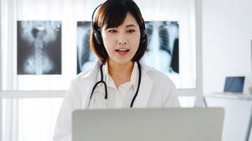 Joven doctora asiática en uniforme médico blanco con estetoscopio usando computadora portátil hablando por videoconferencia con el paciente en el escritorio en la clínica de salud u hospital. concepto de consulta y terapia foto