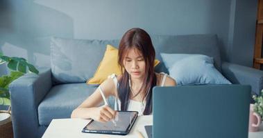Young Asia girl teenager with casual shirt wear earphone use laptop learn online write lecture in laptop in living room at house. Isolate education online e-learning coronavirus pandemic concept. photo