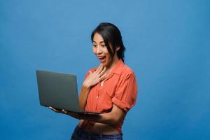 Jovencita asiática usando laptop con expresión positiva, sonríe ampliamente, vestida con ropa casual sintiendo felicidad y parada aislada sobre fondo azul. feliz adorable mujer alegre se regocija con el éxito. foto