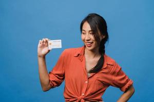 Joven mujer asiática muestra una tarjeta bancaria de crédito con expresión positiva, sonríe ampliamente, vestida con ropa informal sintiendo felicidad y parada aislada sobre fondo azul. concepto de expresión facial. foto