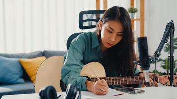 feliz mujer asiática compositora tocar la guitarra acústica escuchar la canción desde el teléfono inteligente pensar y escribir notas letras de canciones en papel sentarse en la sala de estar en casa estudio. producción musical en el concepto de casa. foto
