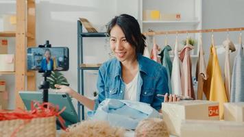 Young Asia lady fashion designer using mobile phone receiving purchase order and showing clothes recording video live streaming online at shop. Small business owner, online market delivery concept. photo