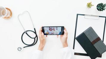 Vista superior de la joven doctora asiática en uniforme médico con estetoscopio usando el teléfono hablando por videoconferencia con el paciente en el escritorio en la clínica de salud u hospital. concepto de consulta y terapia. foto