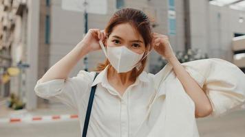 Successful young Asia businesswoman in fashion office clothes wear medical face mask smiling and looking at camera while happy standing alone outdoors in urban modern city. Business on the go concept.