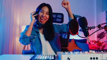 Happy asia girl dj play launchpad synthesizer keyboard sound mixer wear headphone and performance music online live talk with audience in living room home studio at night. Content creator concept.
