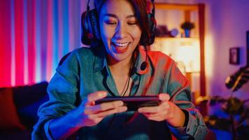 Happy asia girl gamer wear headphone competition play video game online with smartphone colorful neon lights in living room at home. Esport streaming game online, Home quarantine activity concept.