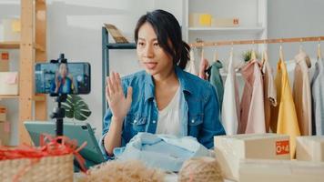 Young Asia lady fashion designer using mobile phone receiving purchase order and showing clothes recording video live streaming online at shop. Small business owner, online market delivery concept. photo