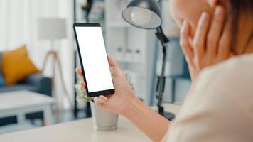 Jovencita asiática usa un teléfono inteligente con una pantalla en blanco en blanco simulado para mostrar texto publicitario mientras trabaja de manera inteligente desde casa en la sala de estar. tecnología chroma key, concepto de diseño de marketing.