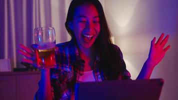 Young Asia lady drinking beer having fun happy moment disco neon night party event online celebration via video call in living room at home. Social distancing, quarantine for coronavirus prevention. photo