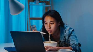 Asia freelance smart business women eating instant noodles while working on laptop in living room at home at night. Happy young Asian girl sitting on desk work overtime, enjoy relax time. photo
