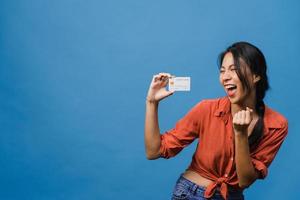 Joven mujer asiática muestra una tarjeta bancaria de crédito con expresión positiva, sonríe ampliamente, vestida con ropa informal sintiendo felicidad y parada aislada sobre fondo azul. concepto de expresión facial. foto